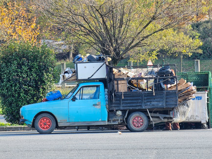 A collection of discarded household waste and packaging cluttering a paved urban area in front of a green metal fence with intricate graffiti artwork in white and light blue. The waste includes a larg
