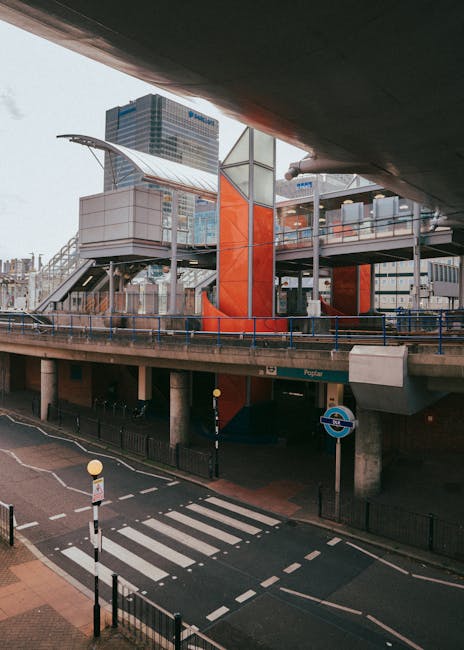 A modern, multi-level pedestrian footbridge with a glass and metal structure, featuring an orange vertical support column, spans across an urban roadway beneath an overpass. The footbridge has a sleek